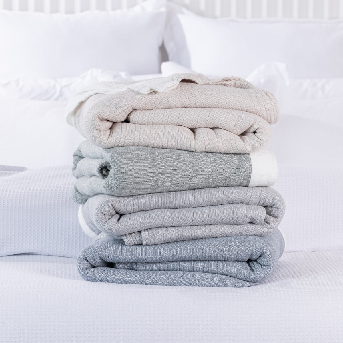 Four folded Cove Bayview Muslin Blankets in Natural, Sage, Grey and Denim stacked on a white bed, showcasing their textured fabric and contrasting white borders.