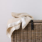 A cream-colored textured wool blanket with white satin trim draped over a brown woven wicker basket against a plain white wall.
