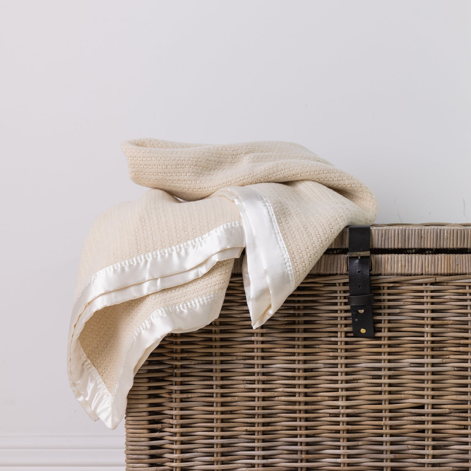 A cream-colored textured wool blanket with white satin trim draped over a brown woven wicker basket against a plain white wall.
