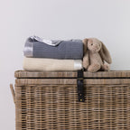 A folded knit blanket in gray and cream are stacked on top of a woven wicker storage chest, with a beige plush bunny toy resting beside them against a plain light gray wall.