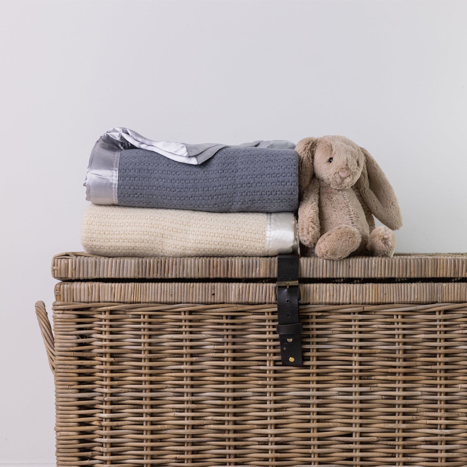 A folded knit blanket in gray and cream are stacked on top of a woven wicker storage chest, with a beige plush bunny toy resting beside them against a plain light gray wall.