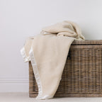 A Cream textured throw blanket with white ruffled trim draped over a wicker storage basket against a plain light gray wall.