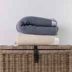 Two folded textured knit blankets, one in slate blue-gray and one in cream, stacked on top of a woven wicker storage basket against a plain light gray wall.