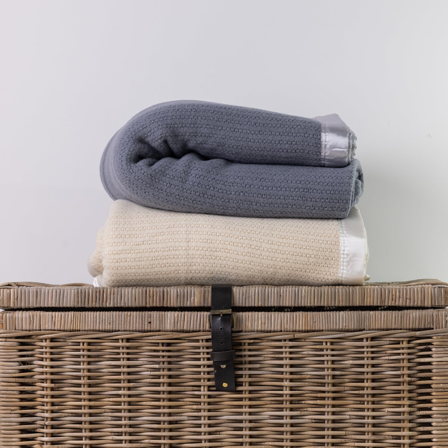 Two folded textured knit blankets, one in slate blue-gray and one in cream, stacked on top of a woven wicker storage basket against a plain light gray wall.