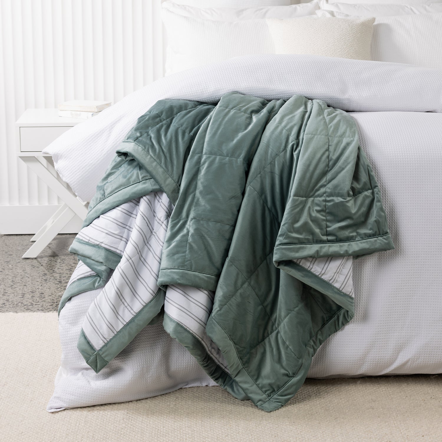 Eucalyptus green quilted blanket draped over the end of a white bed, showing the soft velvet-look front and striped reverse.