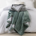 Grey and eucalyptus quilted blankets folded and stacked on a white bed, highlighting the soft velvet-look fabric and striped reverse.