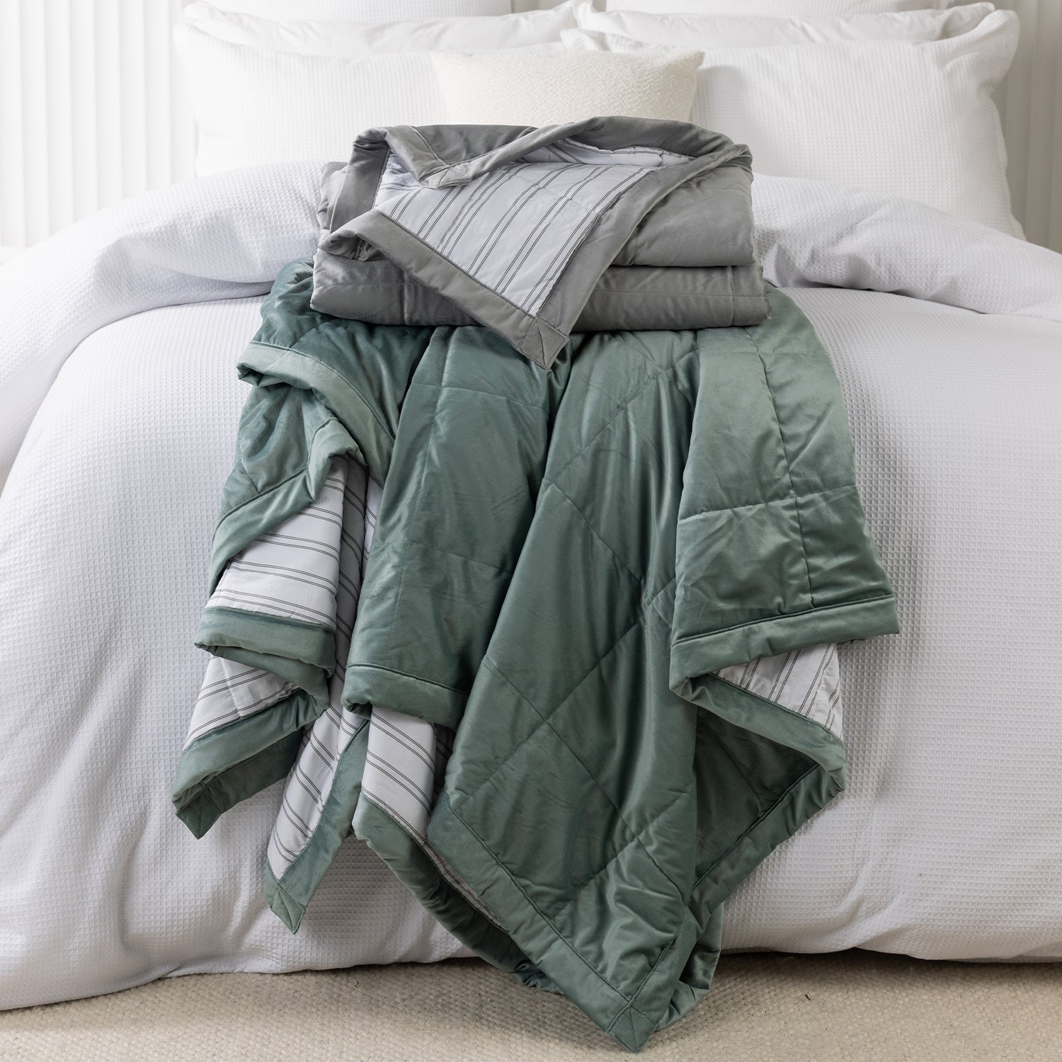Grey and eucalyptus quilted blankets folded and stacked on a white bed, highlighting the soft velvet-look fabric and striped reverse.