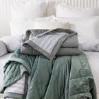 Grey and eucalyptus quilted blankets folded and stacked on a white bed, highlighting the soft velvet-look fabric and striped reverse.