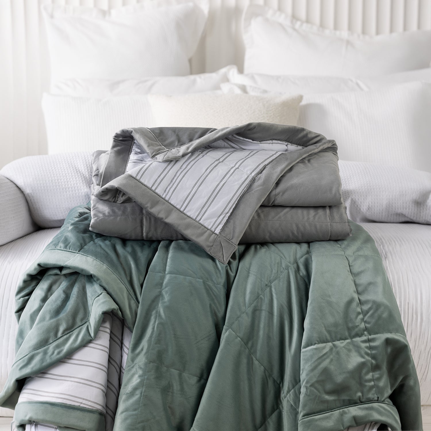 Grey and eucalyptus quilted blankets folded and stacked on a white bed, highlighting the soft velvet-look fabric and striped reverse.