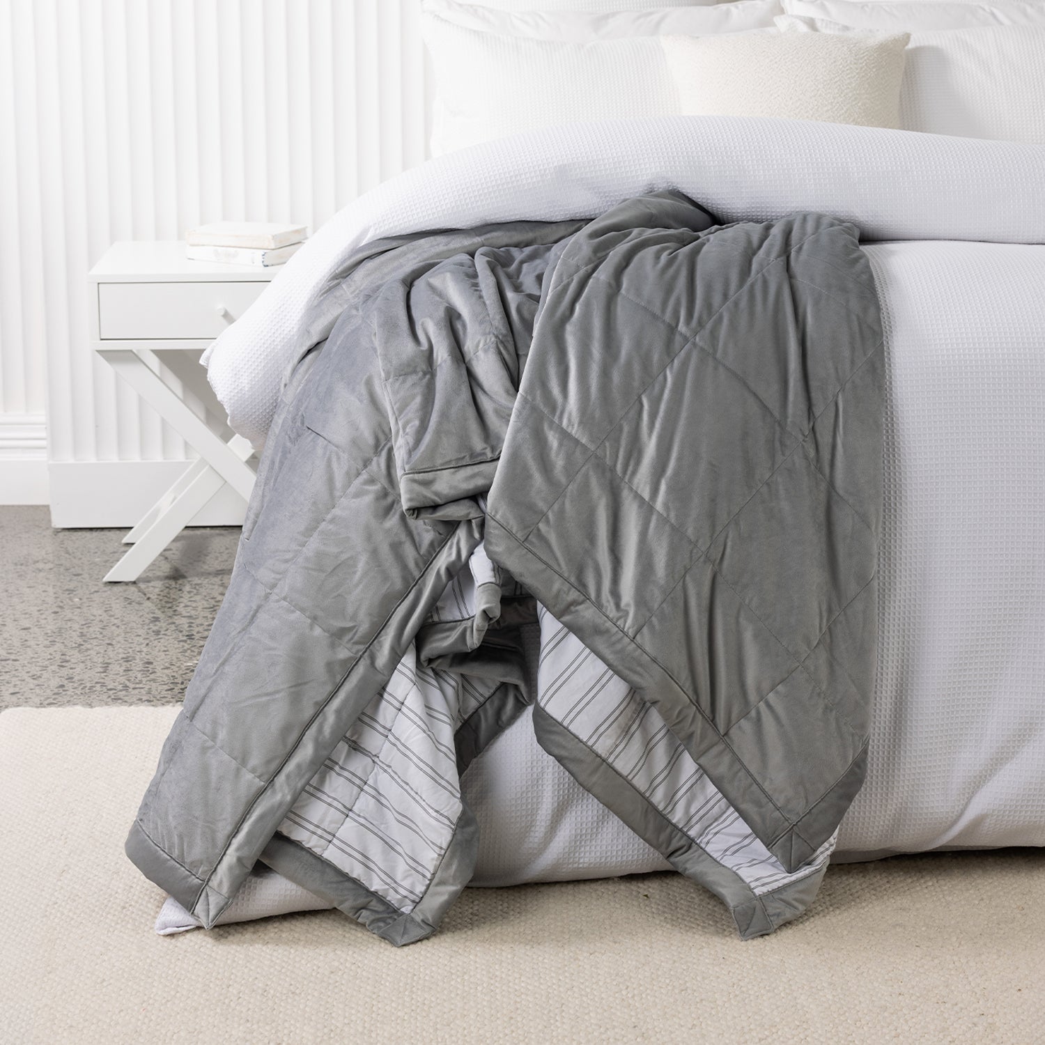 Grey quilted blanket draped over the end of a white bed, highlighting the soft velvet-look fabric and striped reverse.