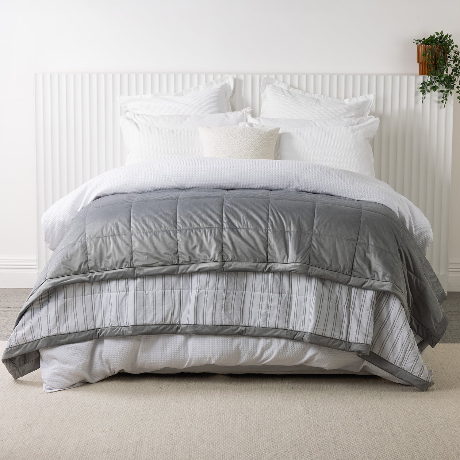 Grey quilted blanket spread across a white bed, displaying the soft velvet-look front with the striped reverse folded over.