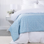 Bambou Summer Blanket in Blue styled on a white bed, featuring a smooth finish and large diamond quilted design.