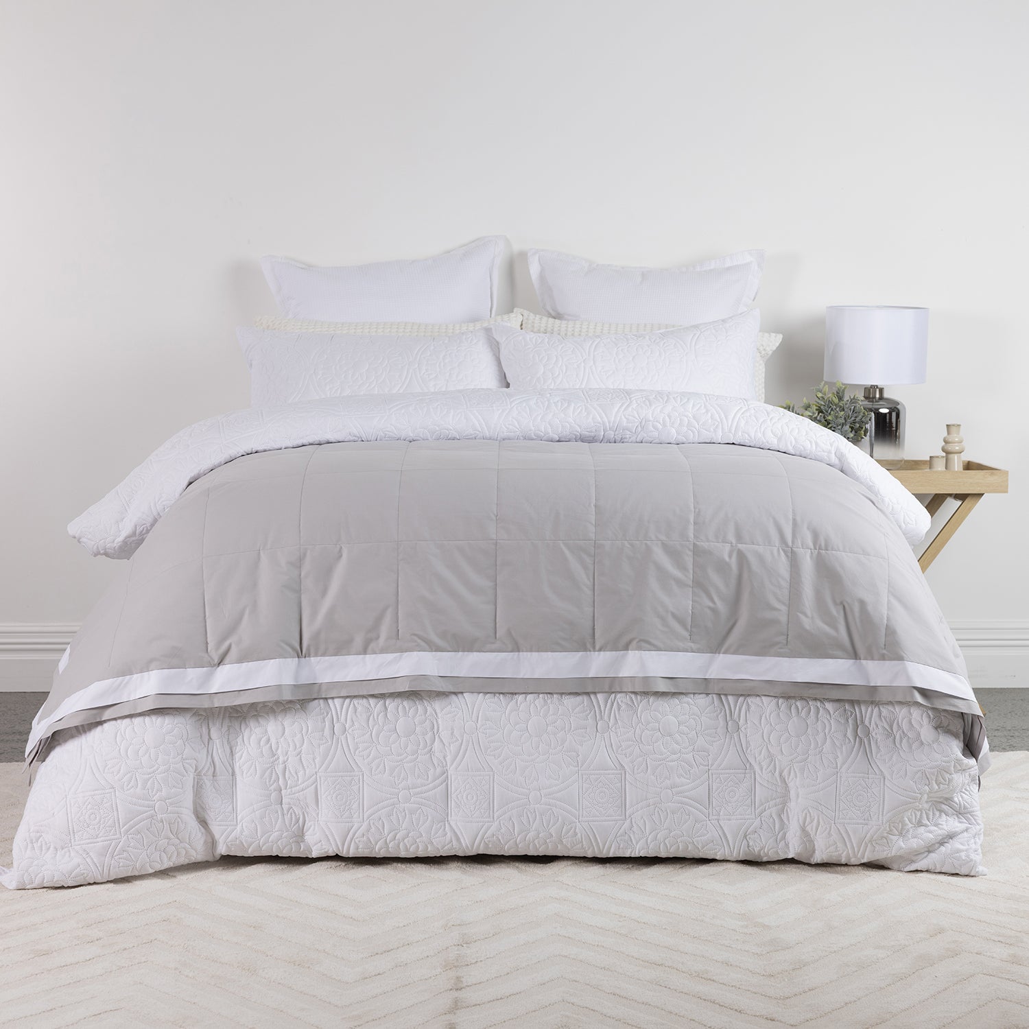 A neatly made bed with a Duck Egg and white quilted comforter set, including matching pillows and a patterned white quilted bed skirt, styled in a bright bedroom setting with a bedside table and lamp.