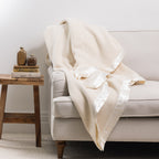 Woolrest 400GSM Wool Blanket in Cream draped over a cream armchair with a satin border, styled beside a wooden side table with books and decorative vases.