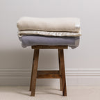 Folded Woolrest 400GSM Wool Blankets in Cream and Glacier displayed on a wooden stool, highlighting the soft wool finish and satin trim.