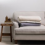 Folded Woolrest 400GSM Wool Blankets in Cream and Glacier stacked on a cream armchair, showcasing the two available colour options.
