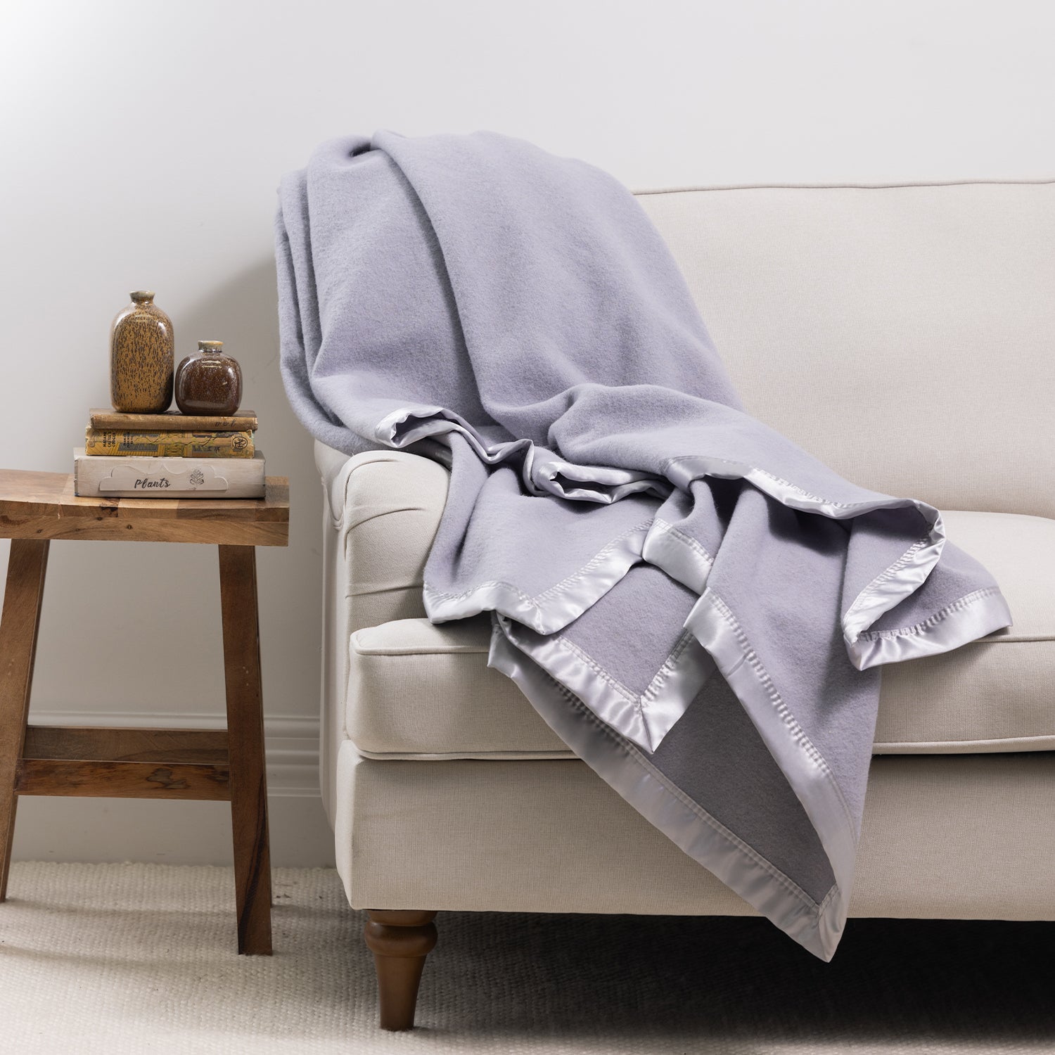 Woolrest 400GSM Wool Blanket in Glacier draped over a cream armchair with a satin border, styled beside a wooden side table with books and decorative vases.