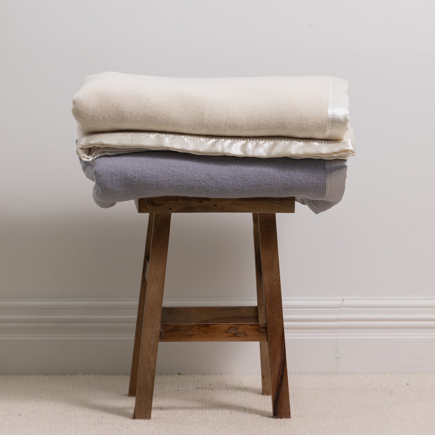 Folded Woolrest 400GSM Wool Blankets in Cream and Glacier displayed on a wooden stool, highlighting the soft wool finish and satin trim.