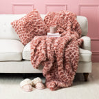 Pink faux fur blanket and cushion set styled on a cream sofa with pink slippers and a mug.