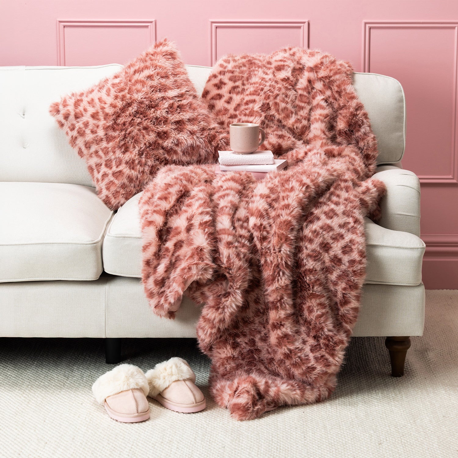 Pink faux fur blanket and cushion set styled on a cream sofa with pink slippers and a mug.