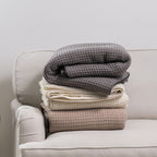 Stack of Charcoal, Ivory, and Moonlight waffle weave blankets folded on a cream sofa, showcasing the textured waffle finish in each colour.