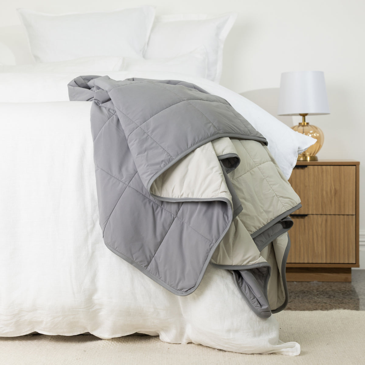 Grey quilted cooling blanket draped over the edge of a white bed, showing its soft padded construction, square stitching and contrasting lighter reverse.