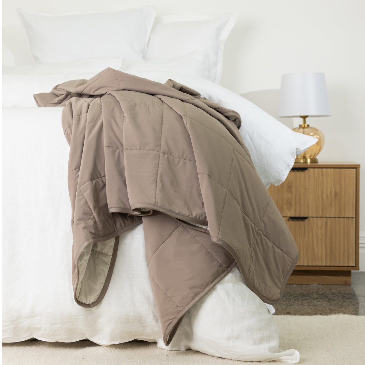 Mocha quilted cooling blanket casually draped over a white bed, highlighting the smooth fabric, padded square quilting and bound edges.