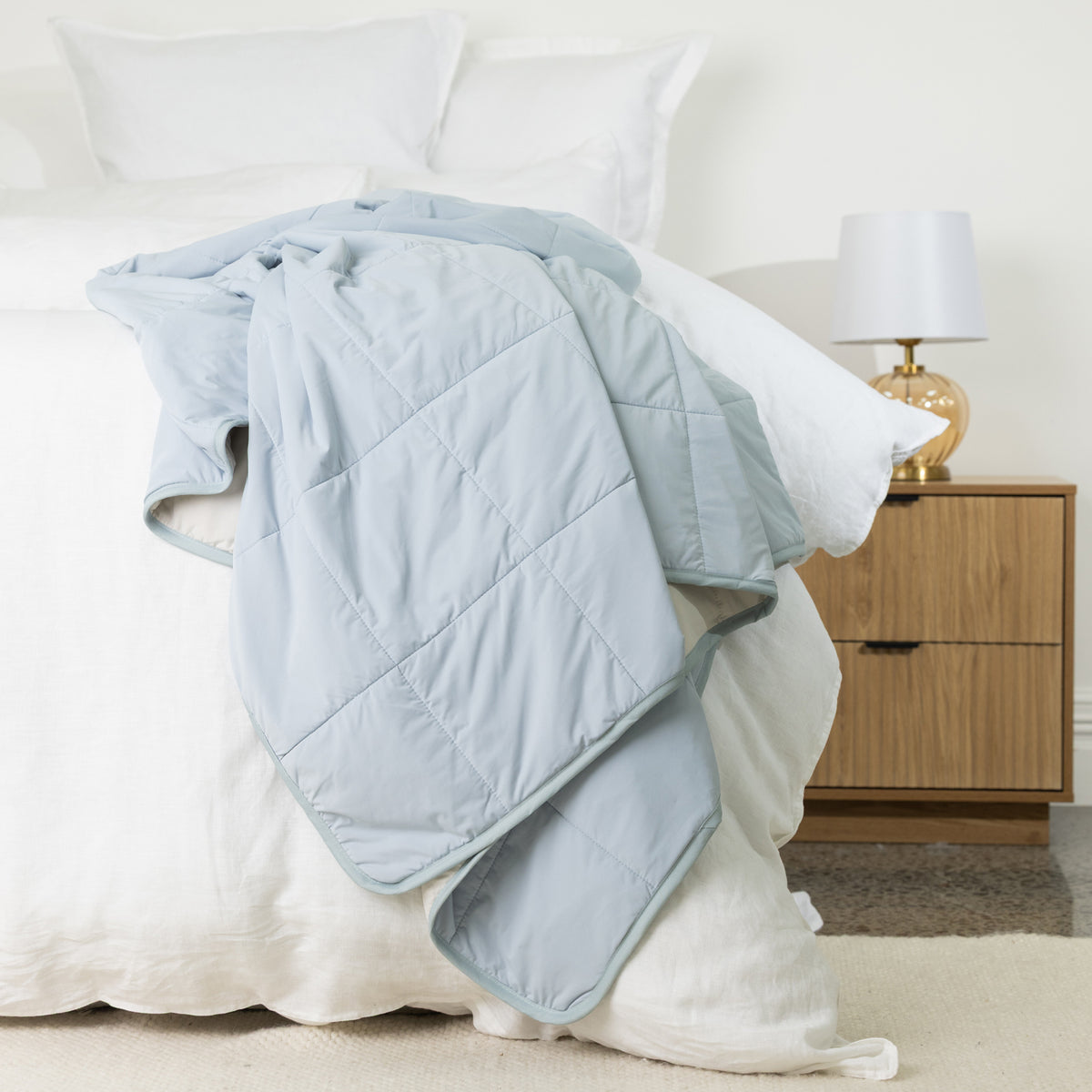Pale blue quilted cooling blanket displayed over the side of a white bed, showing its soft lightweight padding, square stitched design and neat piped edging.
