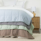 Close-up of four quilted cooling blankets layered over the edge of a white bed in pale blue, grey, seafoam and mocha, showing the stitched construction and colour range.