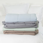 Folded quilted cooling blankets stacked on a white bed in pale blue, grey, seafoam and mocha, displaying the coordinated colour options and padded stitched design.