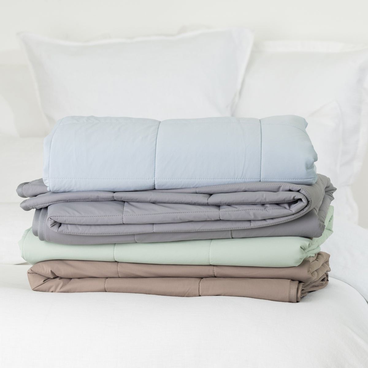 Folded quilted cooling blankets stacked on a white bed in pale blue, grey, seafoam and mocha, displaying the coordinated colour options and padded stitched design.