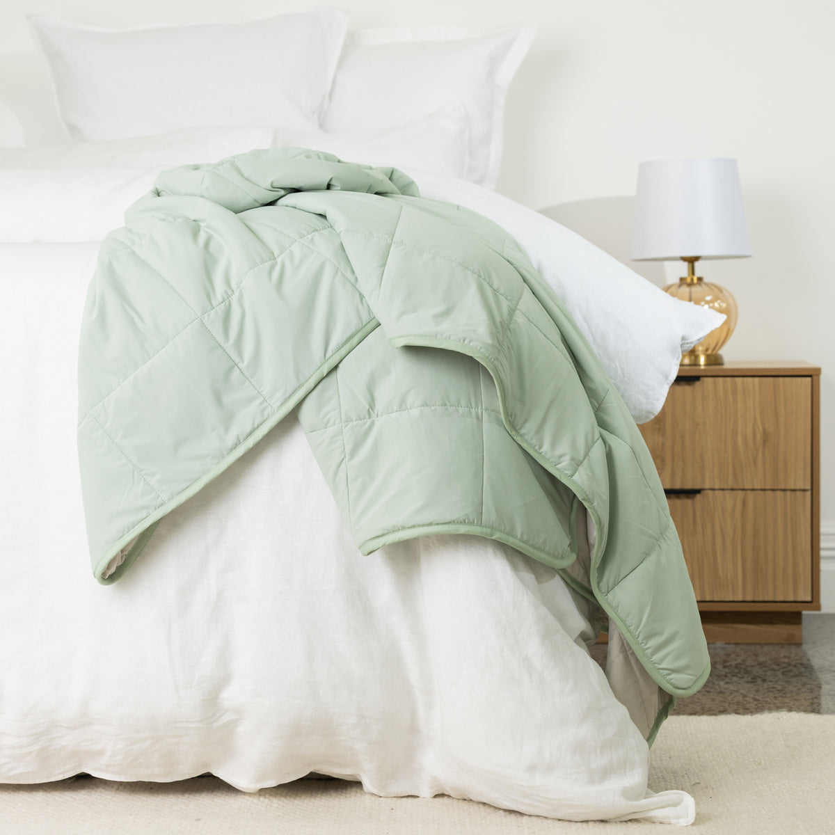 Seafoam quilted cooling blanket draped across a white bed, featuring a soft pastel green finish, square quilting and smooth bound edges.