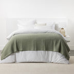 Sage green stonewashed cotton blanket layered across a white bed with crisp white bedding and a matching bedside table.