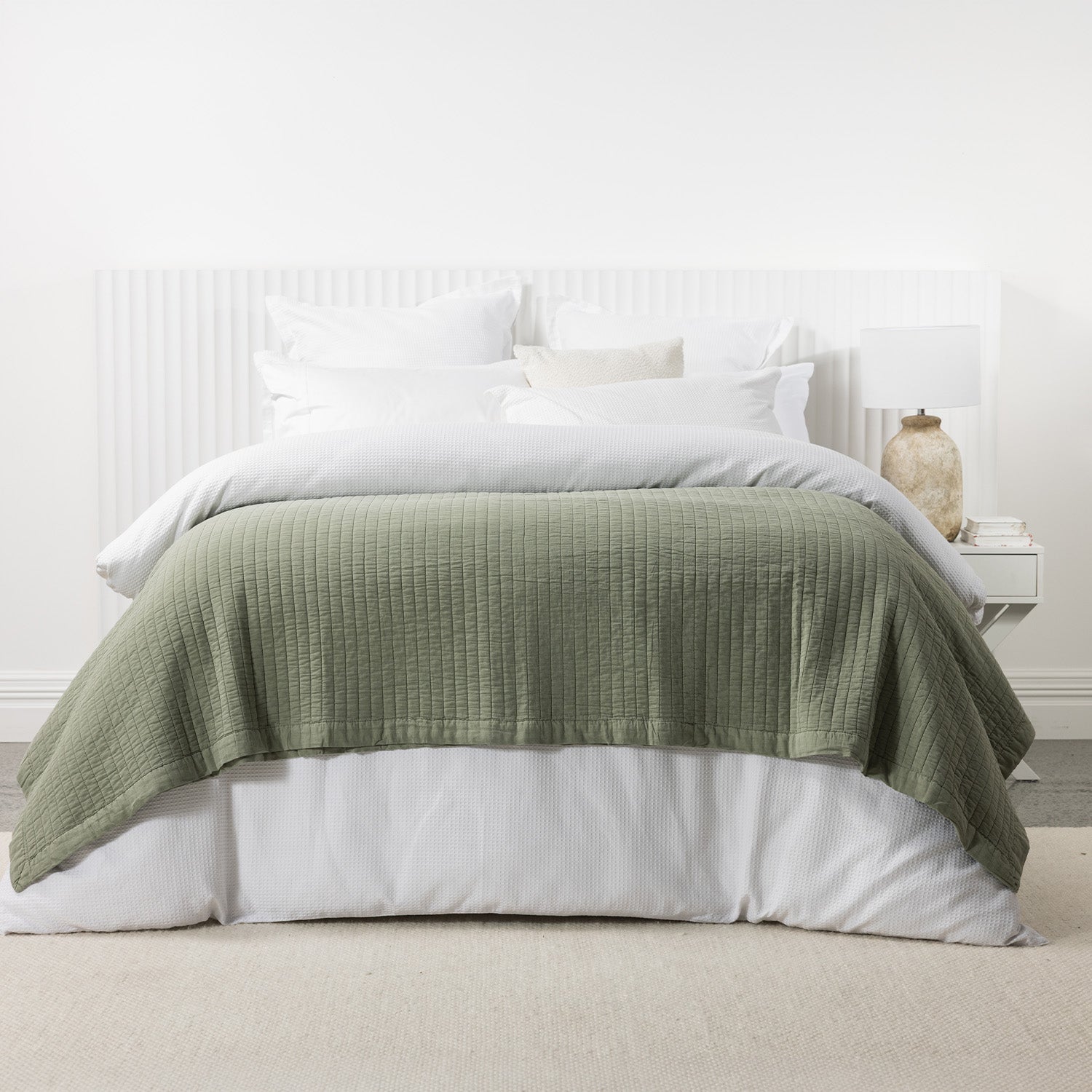 Sage green stonewashed cotton blanket layered across a white bed with crisp white bedding and a matching bedside table.