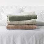 Folded Moss, Natural, and Pecan stonewashed cotton blankets stacked on a white bed, showcasing the quilted texture and available colourways.