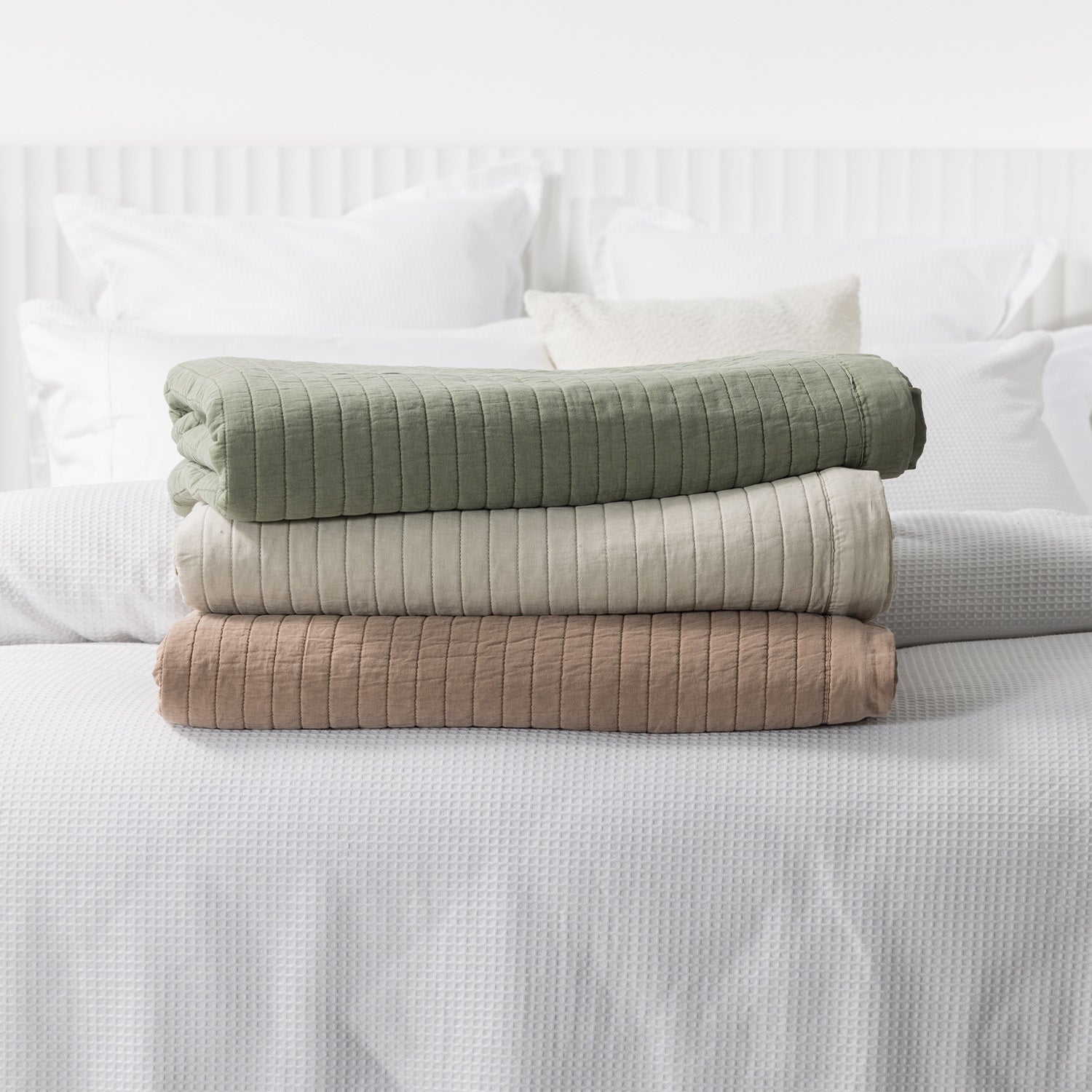 Folded Moss, Natural, and Pecan stonewashed cotton blankets stacked on a white bed, showcasing the quilted texture and available colourways.