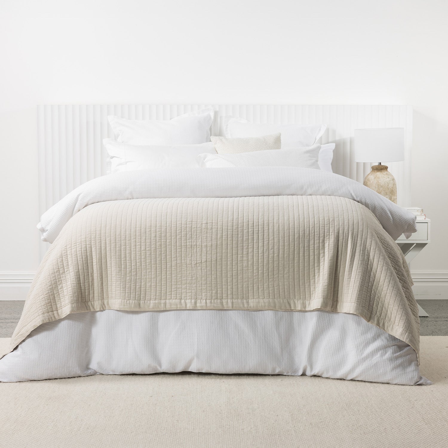 Natural stonewashed cotton blanket layered across a white bed with crisp white bedding and a matching bedside table.