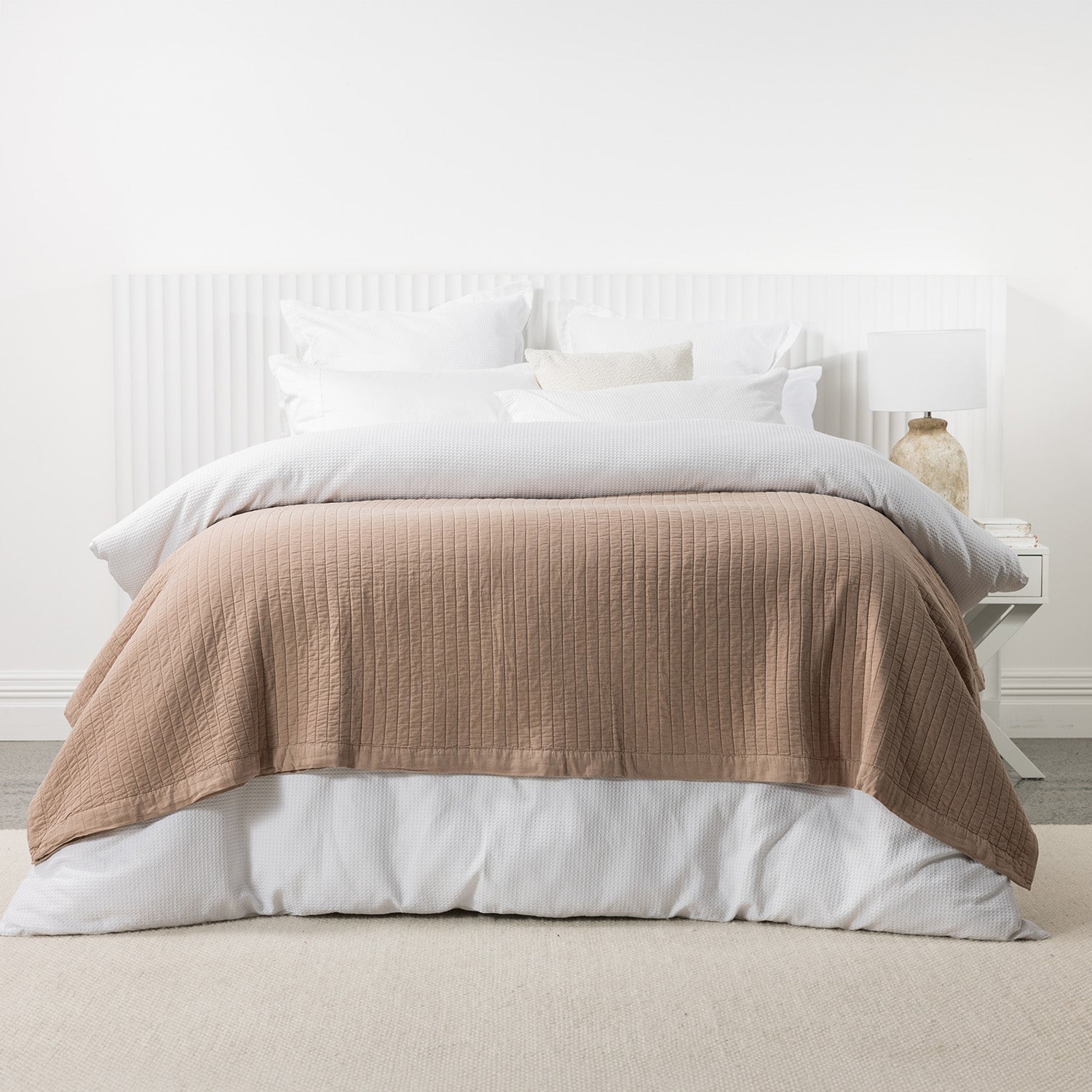 Pecan stonewashed cotton blanket layered across a white bed with crisp white bedding and a matching bedside table.