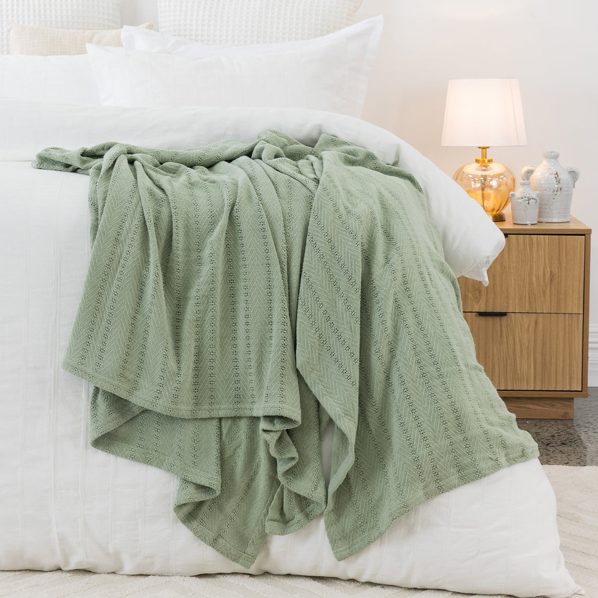 Sage aircell blanket draped across a white bed, showcasing its lightweight textured knit and subtle striped pattern.