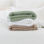 Sage, White and Taupe aircell blankets folded and stacked together on a white bed, showcasing the three colour options.