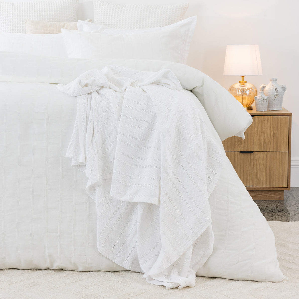 White aircell blanket draped across a white bed, showcasing its lightweight textured knit and subtle striped pattern.