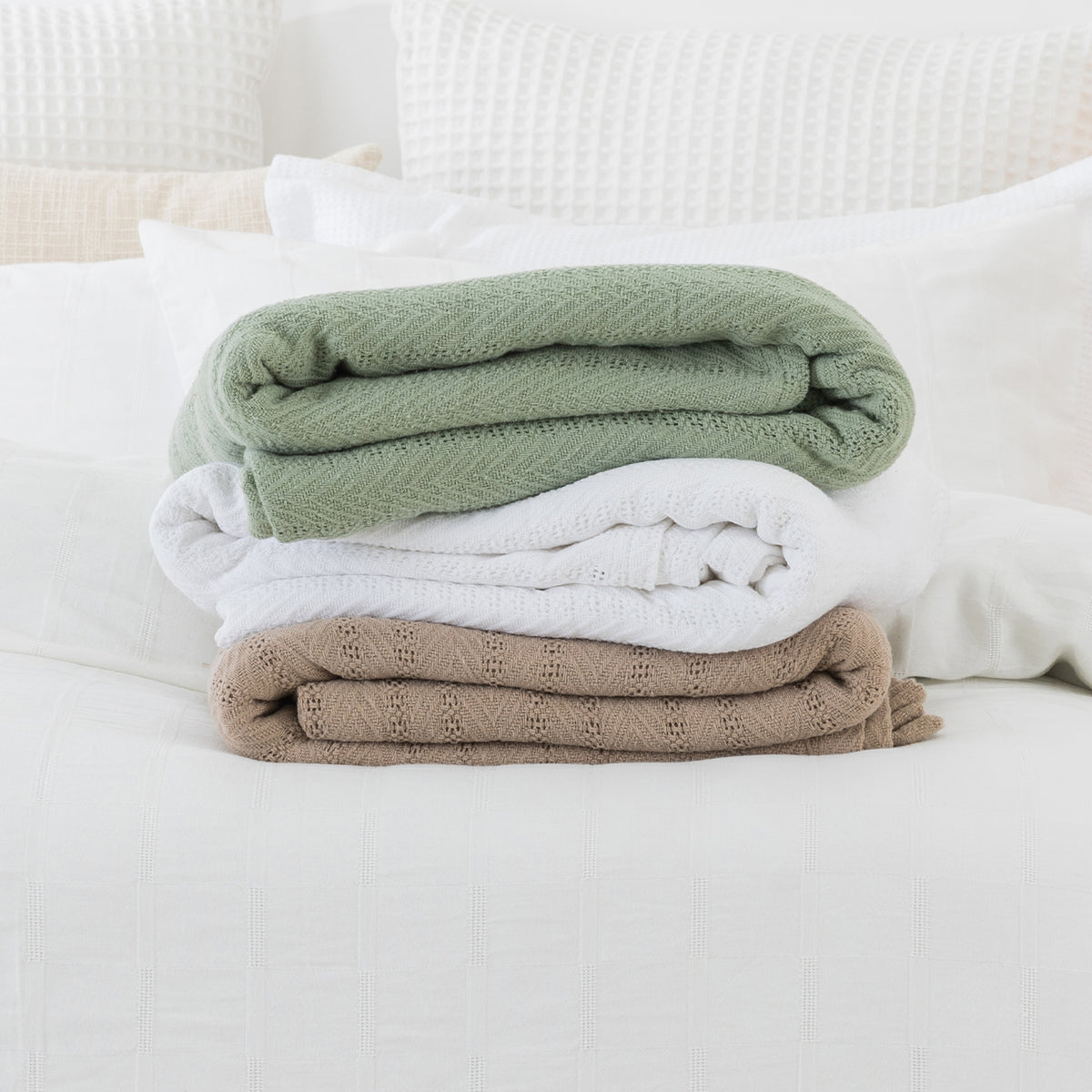 Sage, White and Taupe aircell blankets folded and stacked together on a white bed, showcasing the three colour options.