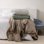 Fieldcrest duck down blankets in Pumice, Moss, and Driftwood, neatly stacked on the end of a white bed, with the Pumice blanket draped in front to showcase the quilted design and satin trim.