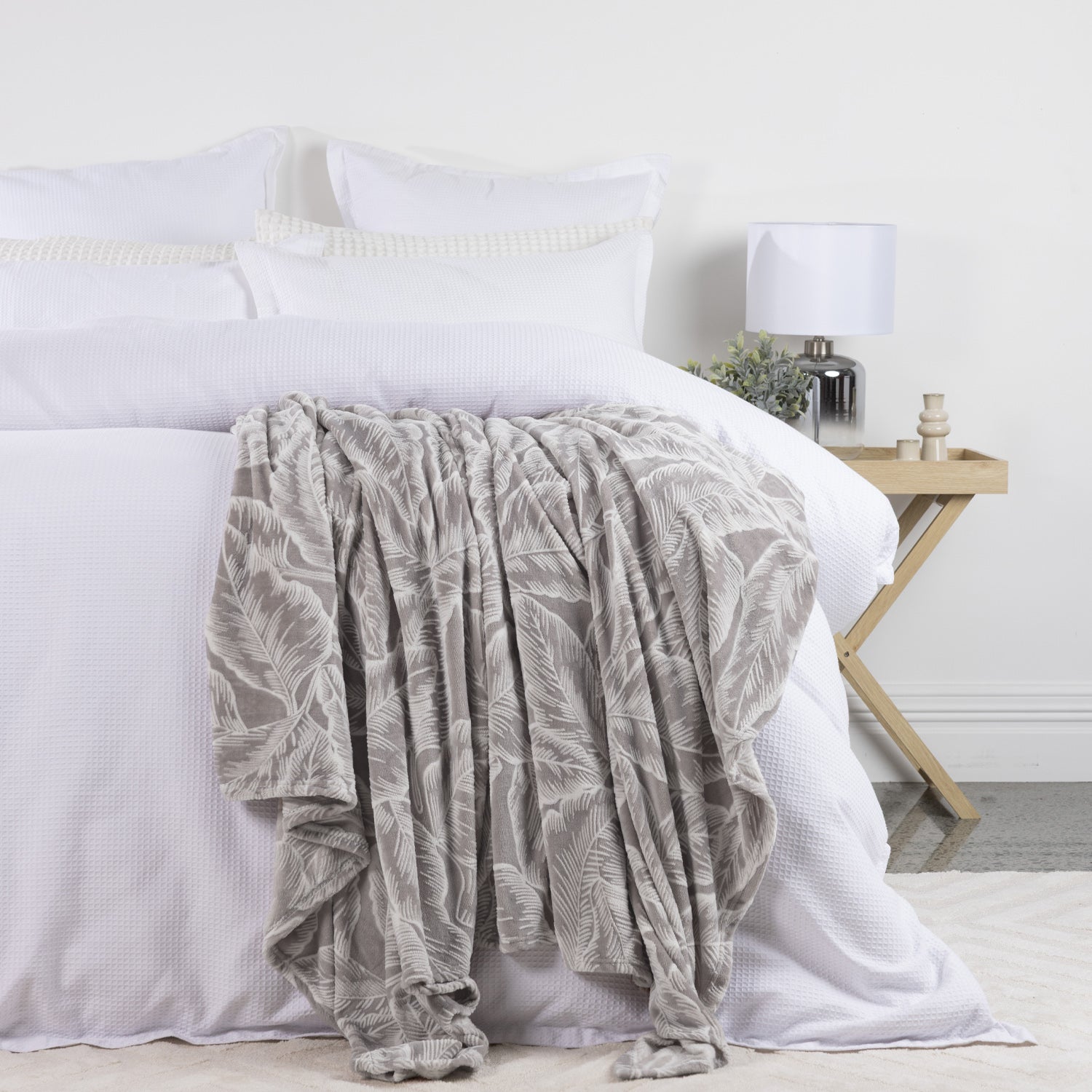 Solace Palmer Blanket in Grey draped across a white bed, featuring a soft botanical leaf pattern in a bright neutral bedroom.