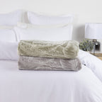 Solace Palmer Blankets in Sage and Grey folded and stacked on a white bed, showing the coordinating botanical leaf design.