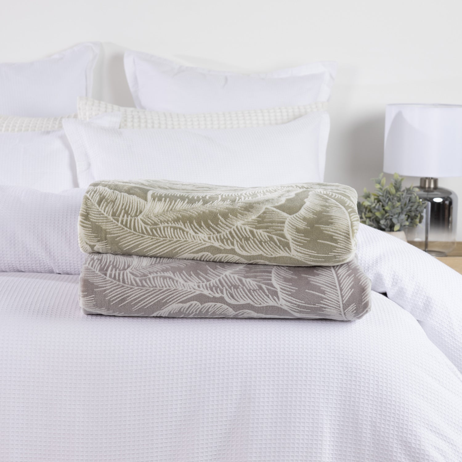 Solace Palmer Blankets in Sage and Grey folded and stacked on a white bed, showing the coordinating botanical leaf design.
