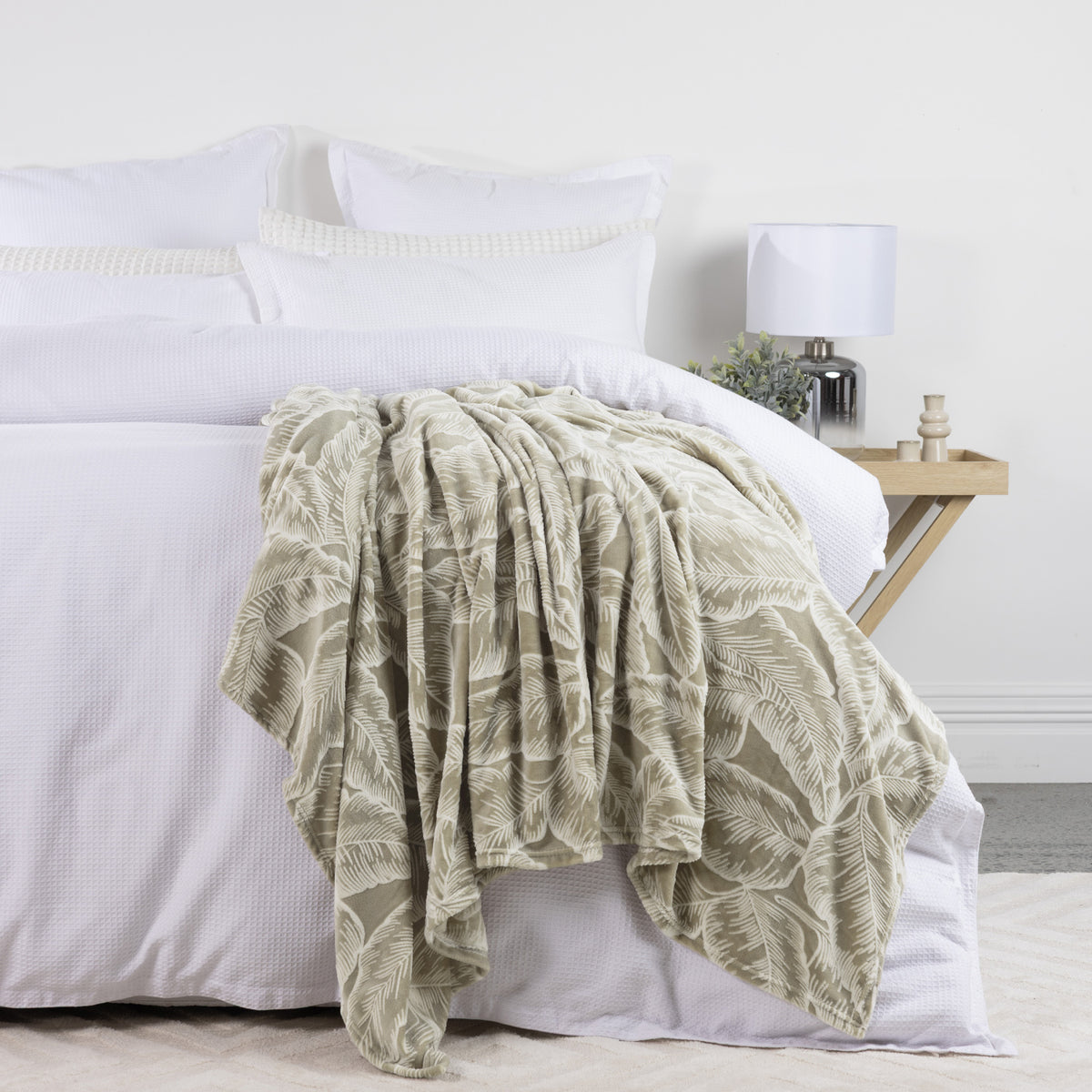 Solace Palmer Blanket in Sage draped across a white bed, showcasing its soft texture and botanical leaf pattern.