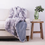 A plush faux-fur throw blanket in a dusty lavender-gray color is draped over a light beige sofa, with a solid blue underside visible where the blanket folds. The scene is styled in a living room setting with a wooden side table holding a green glass vase with a plant, against a clean white wall.