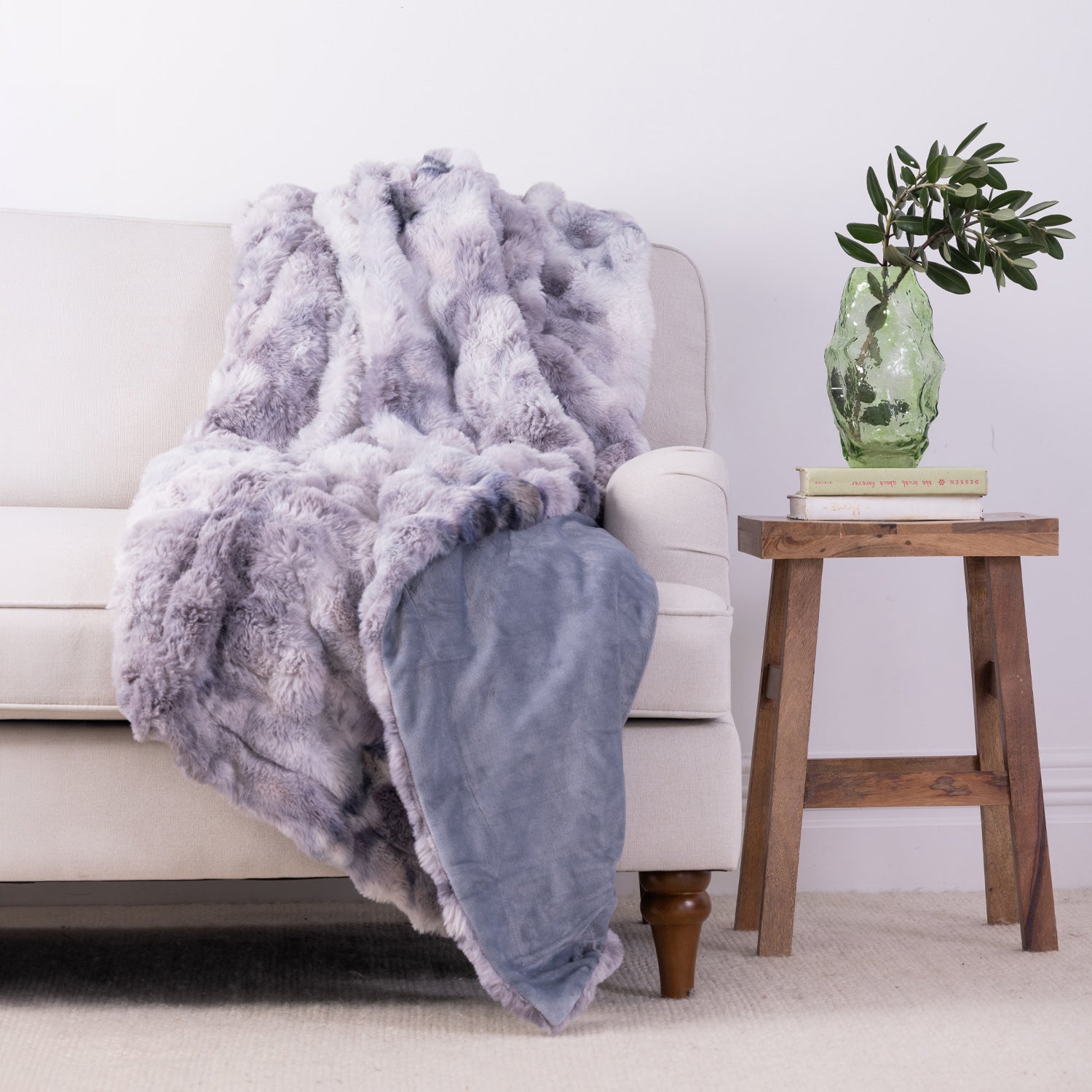A plush faux-fur throw blanket in a dusty lavender-gray color is draped over a light beige sofa, with a solid blue underside visible where the blanket folds. The scene is styled in a living room setting with a wooden side table holding a green glass vase with a plant, against a clean white wall.