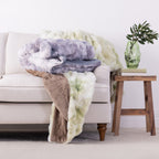 Three soft faux-fur style throw blankets in lavender, cream, and taupe draped over a light beige sofa, with a small wooden side table holding a green glass vase and leaves beside it.